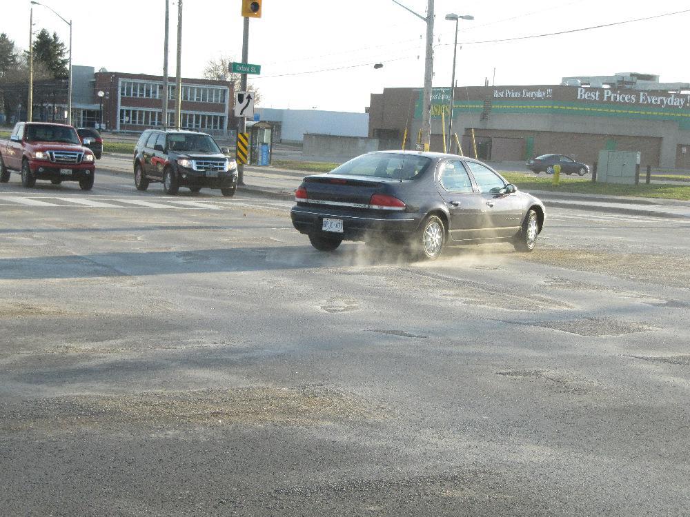 Left-turning vehicle at the intersection of Oxford and Highbury, spins its tires on the loose sand deposited throughout the intersection.